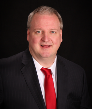 A professional headshot of a man with short, light-colored hair, wearing a dark pinstripe suit, a white dress shirt, and a bold red necktie against a solid black background.