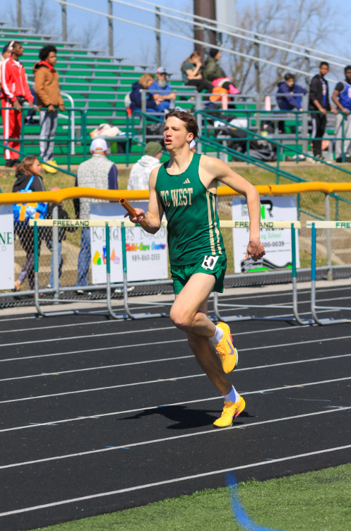 A male track athlete from TC West competes in a relay race on an outdoor black track. He is wearing a green and gold 