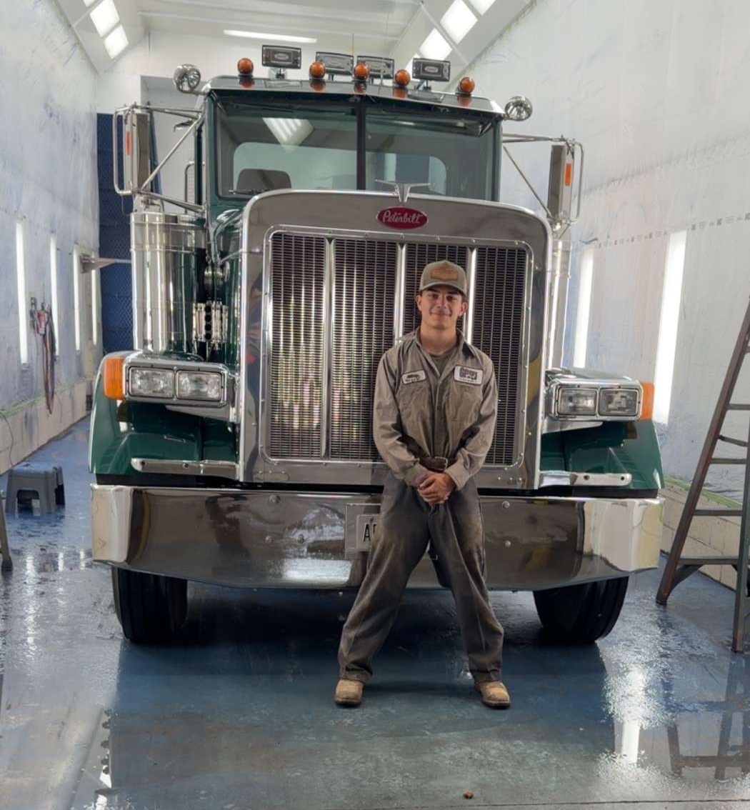 Male wearing grey had and hoodie, standing in front of a tractor trailer