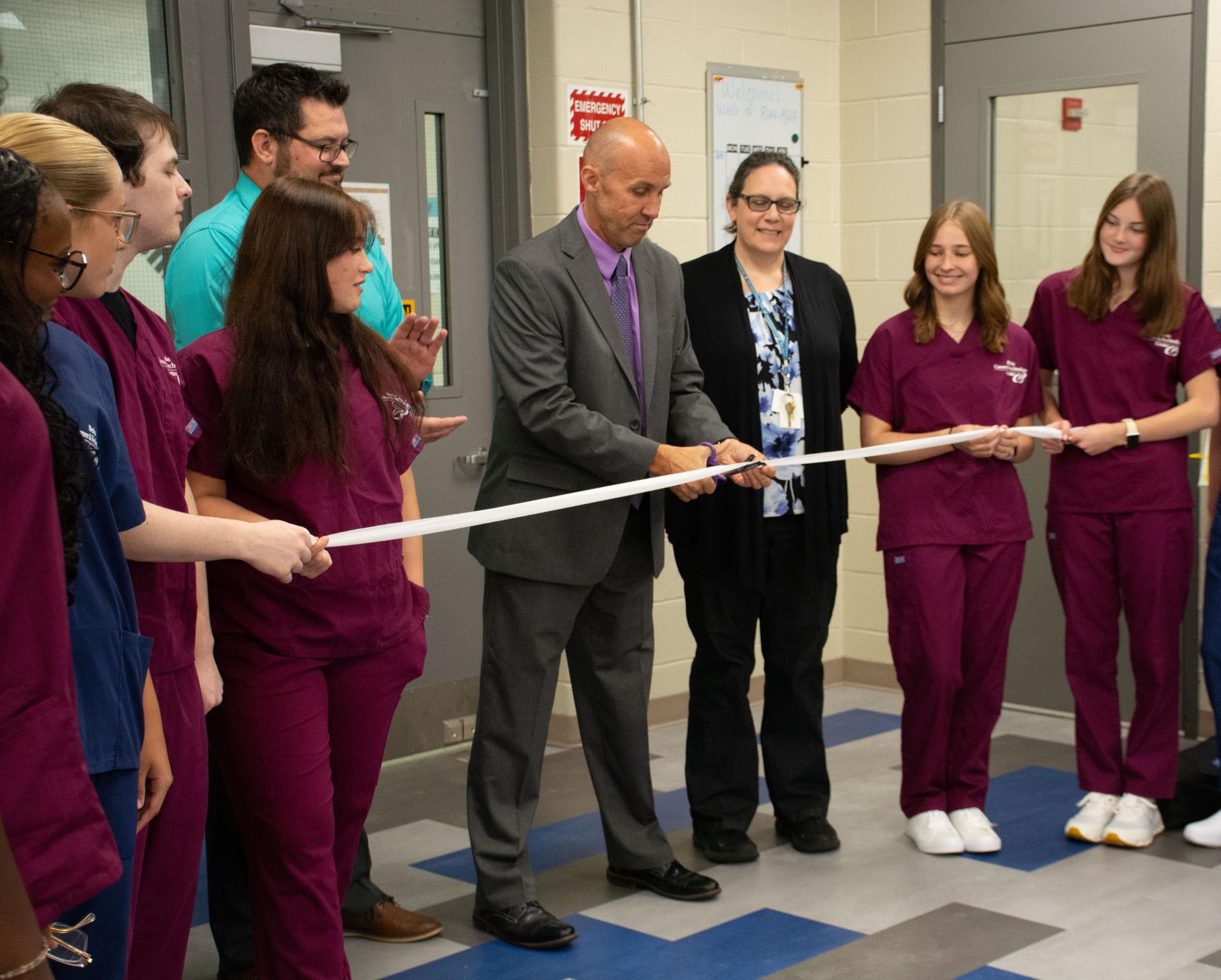 Man standing in grey suit cutting gauze, with a man and woman on each side.  Students wearing maroon scrubs are on each side of adults.