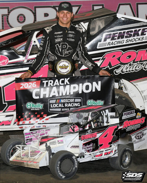 Male in a black cap and black and grey racing gear, holding a 2026 track champion banner and his race car below