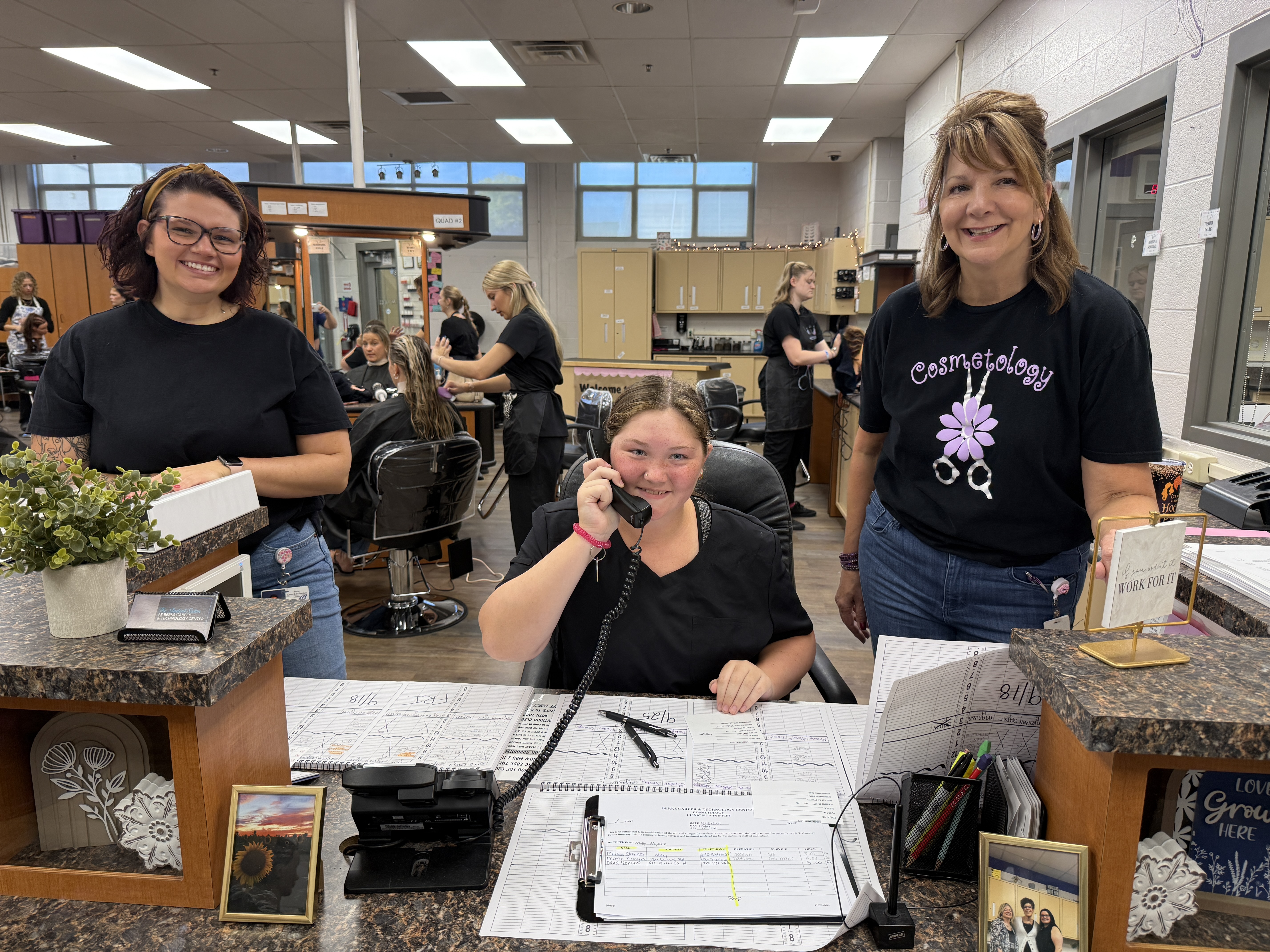 Three females at the front desk of a salon.  Middle female is sitting and on the phone