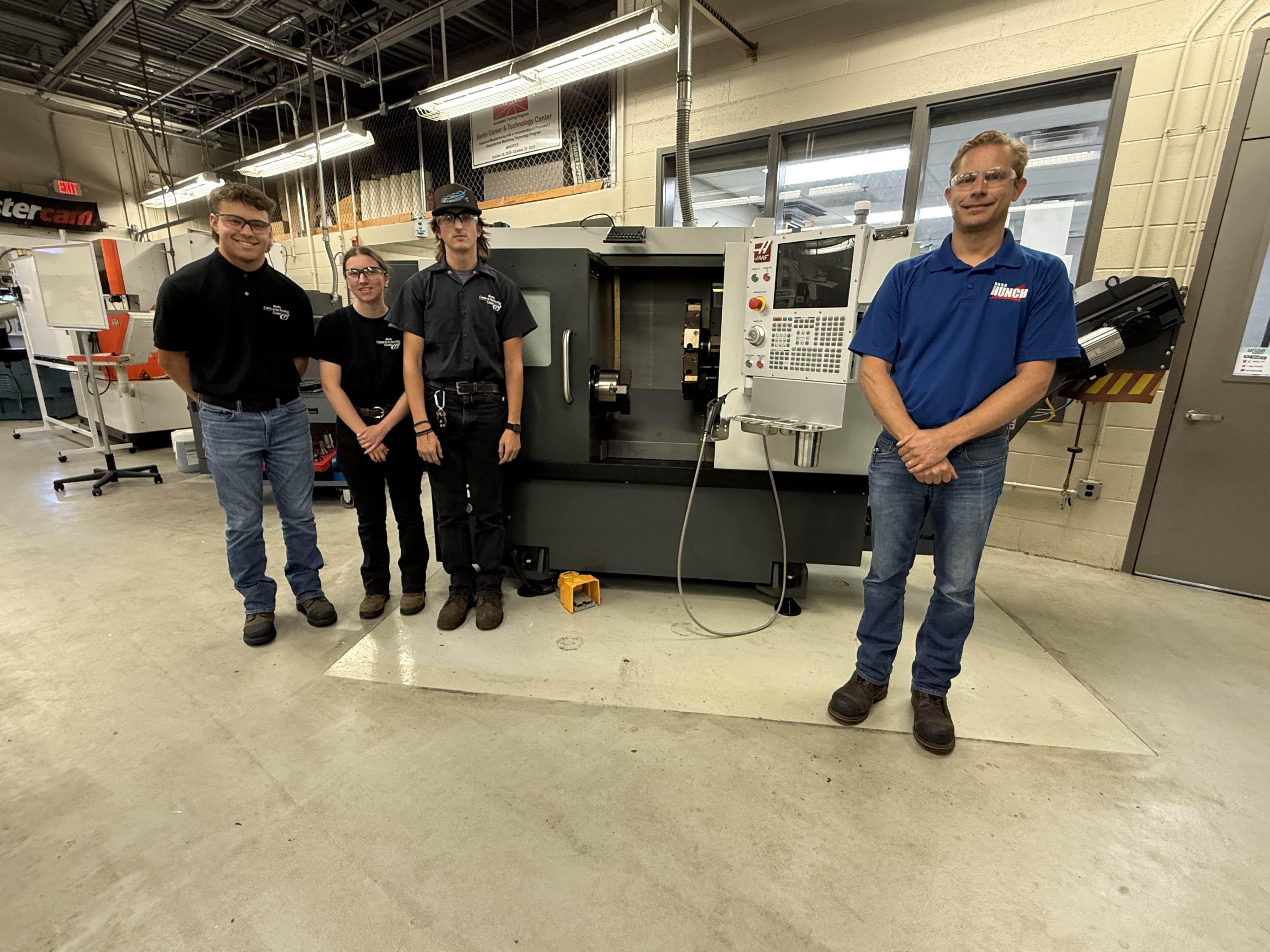 Three students in black shirts and jeans and a male adult in a blue shirt and jeans standing in front of a HAAS computerized lathe.  