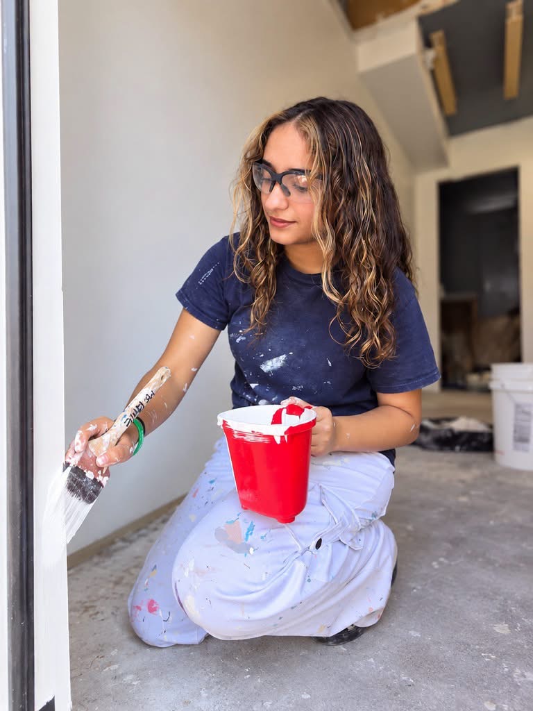 Female wearing safety glasses, navy shirt and white pants painting a wall