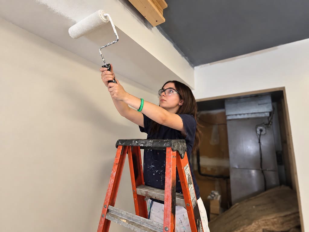 Female on a ladder wearing safety glasses and navy shirt painting a ceiling