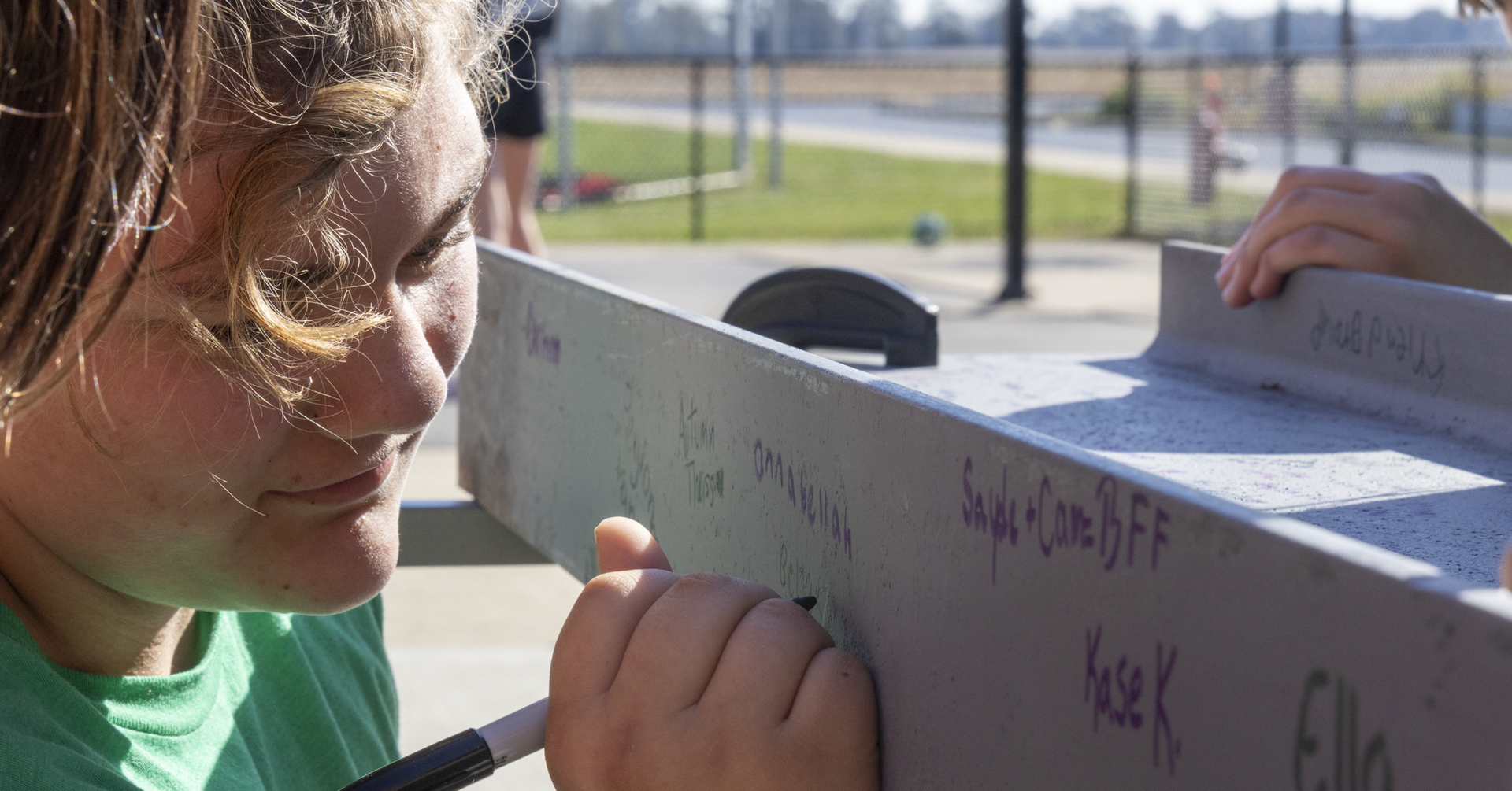 Girl with pen, writing on a tall, rectangular board, with another person's hand touching the board.