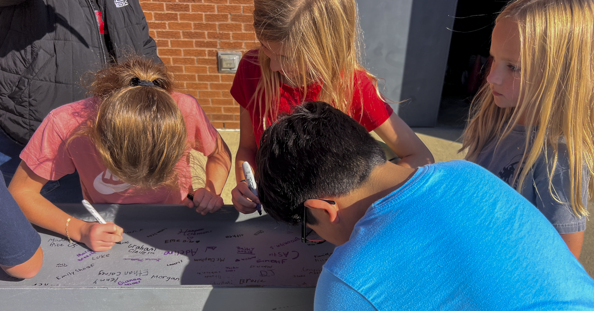 Students sign a large white sheet placed on a table; some have markers in hand.
