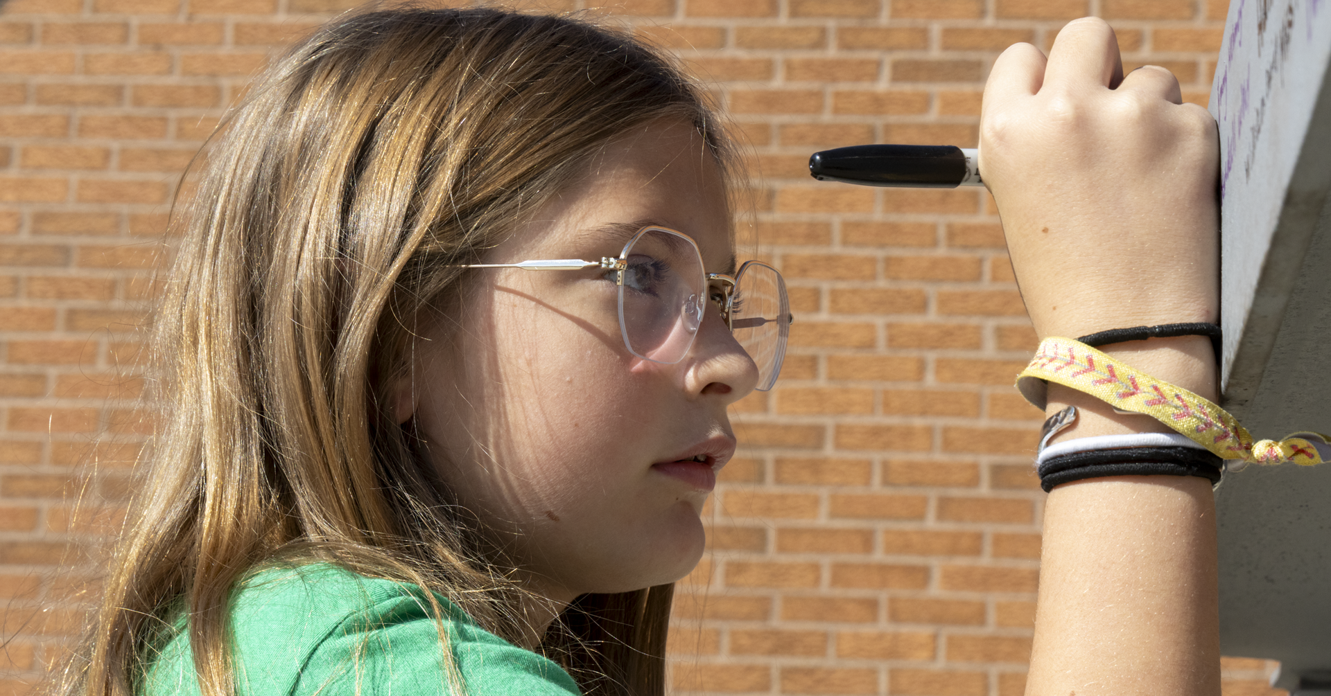 A girl writes on a whiteboard using a black marker, standing in front of a brick wall.