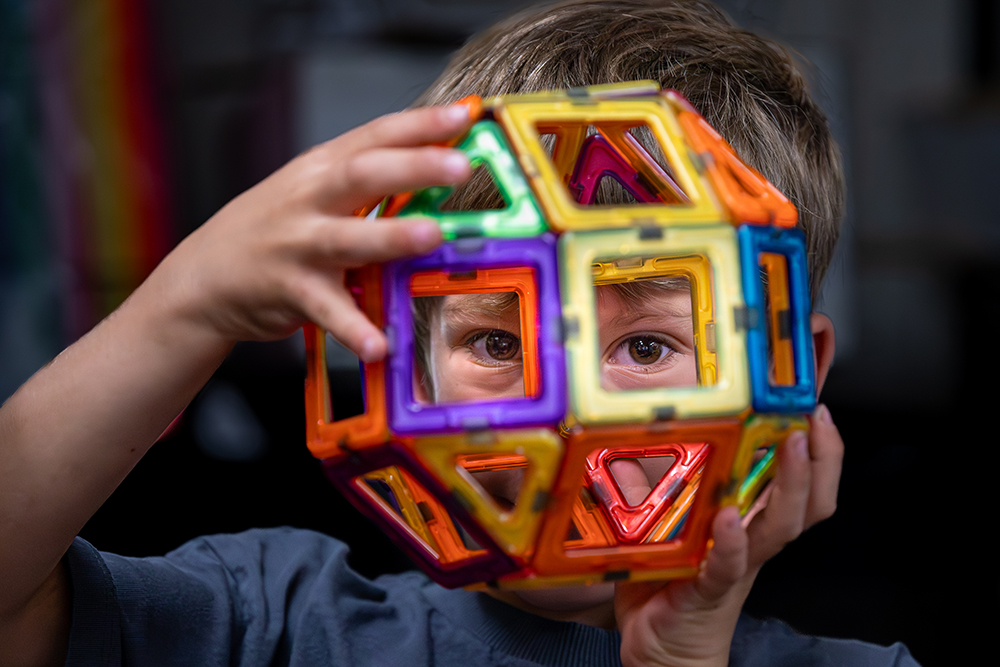 A young student looks through the center of a colorful, geometric sphere built with magnetic tiles.