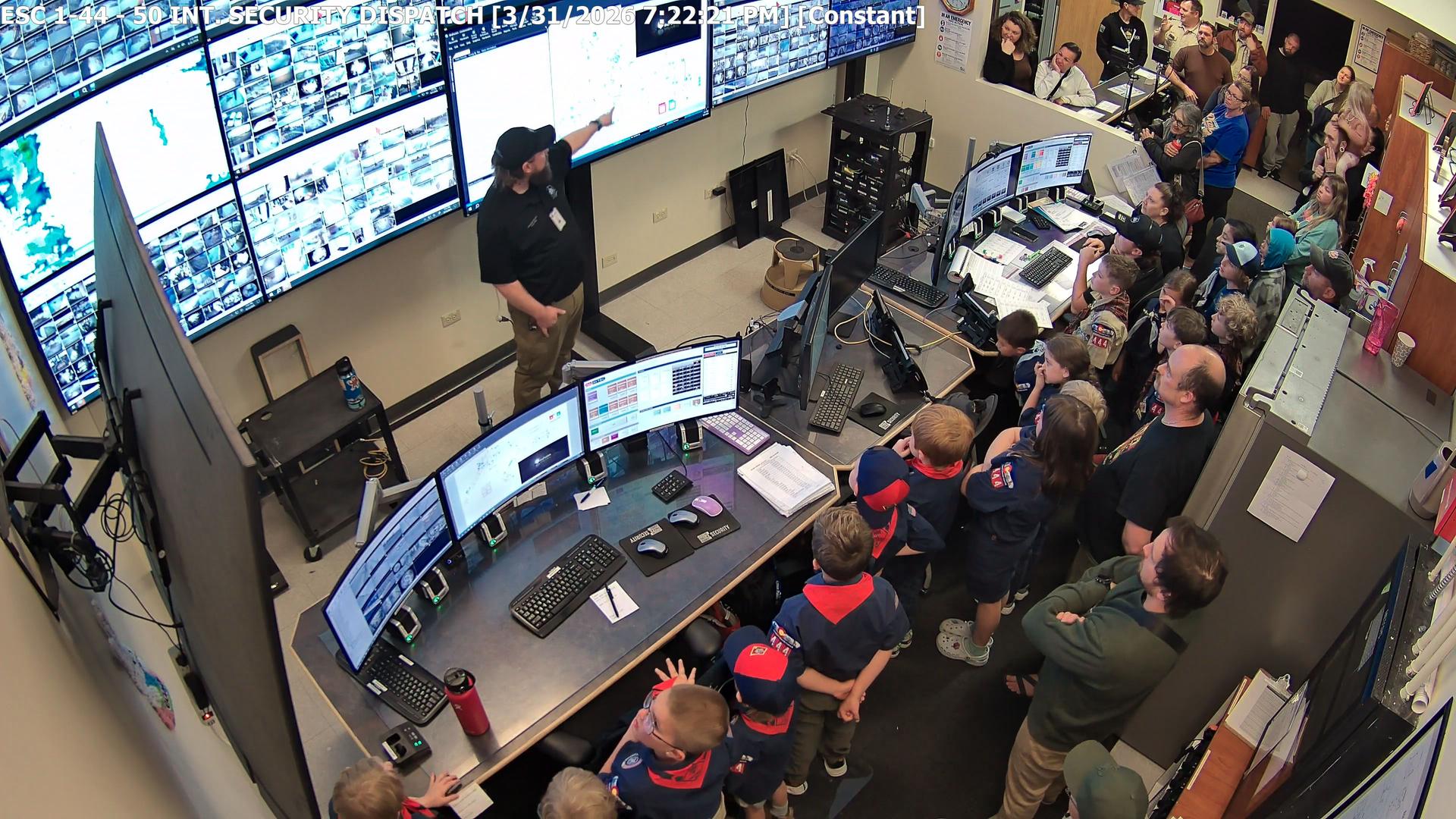 A group of Scouts visits a security dispatch room, watching an officer point to a wall of surveillance monitors.