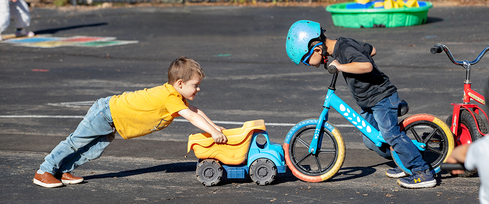 Two children play on a paved playground; one pushes a toy dump truck into the front wheel of another's blue balance bike.