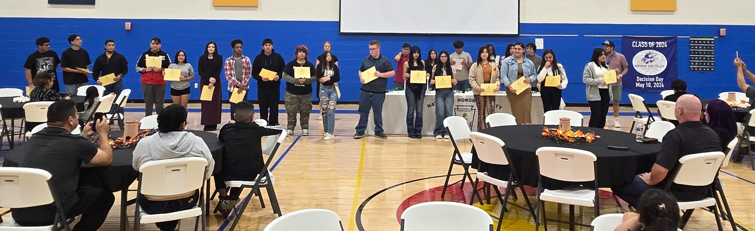 Students standing at the front of the gym in front of tables of parents. Students are holding certificates.