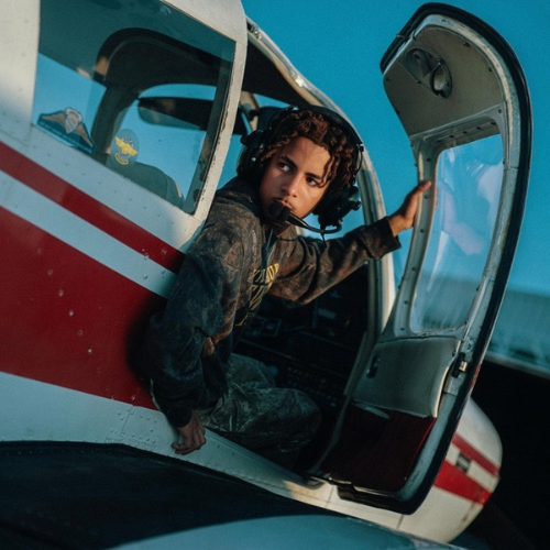 A student wearing a headset sits in the open doorway of a small red and white airplane, looking outside while holding onto the doorframe.