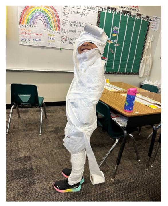 A student wrapped in toilet paper as a mummy smiles to the camera.