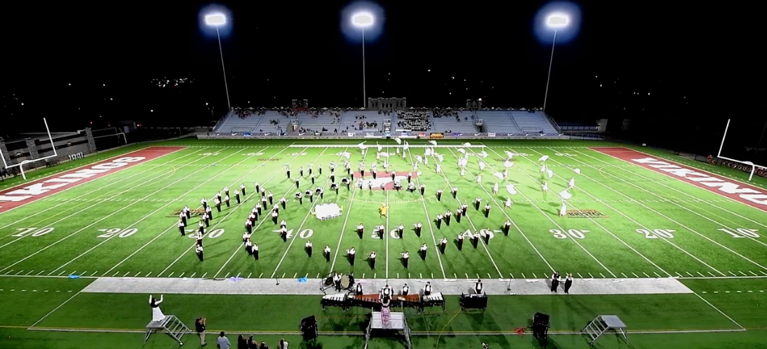 The Tennessee High School Mighty Viking Band performs on the field at night under bright stadium lights during a marching band competition. The band forms a large “V” formation at the 50-yard line while color guard members with white flags are spread across the field. The performance takes place at Stone Castle Stadium with spectators visible in the stands.