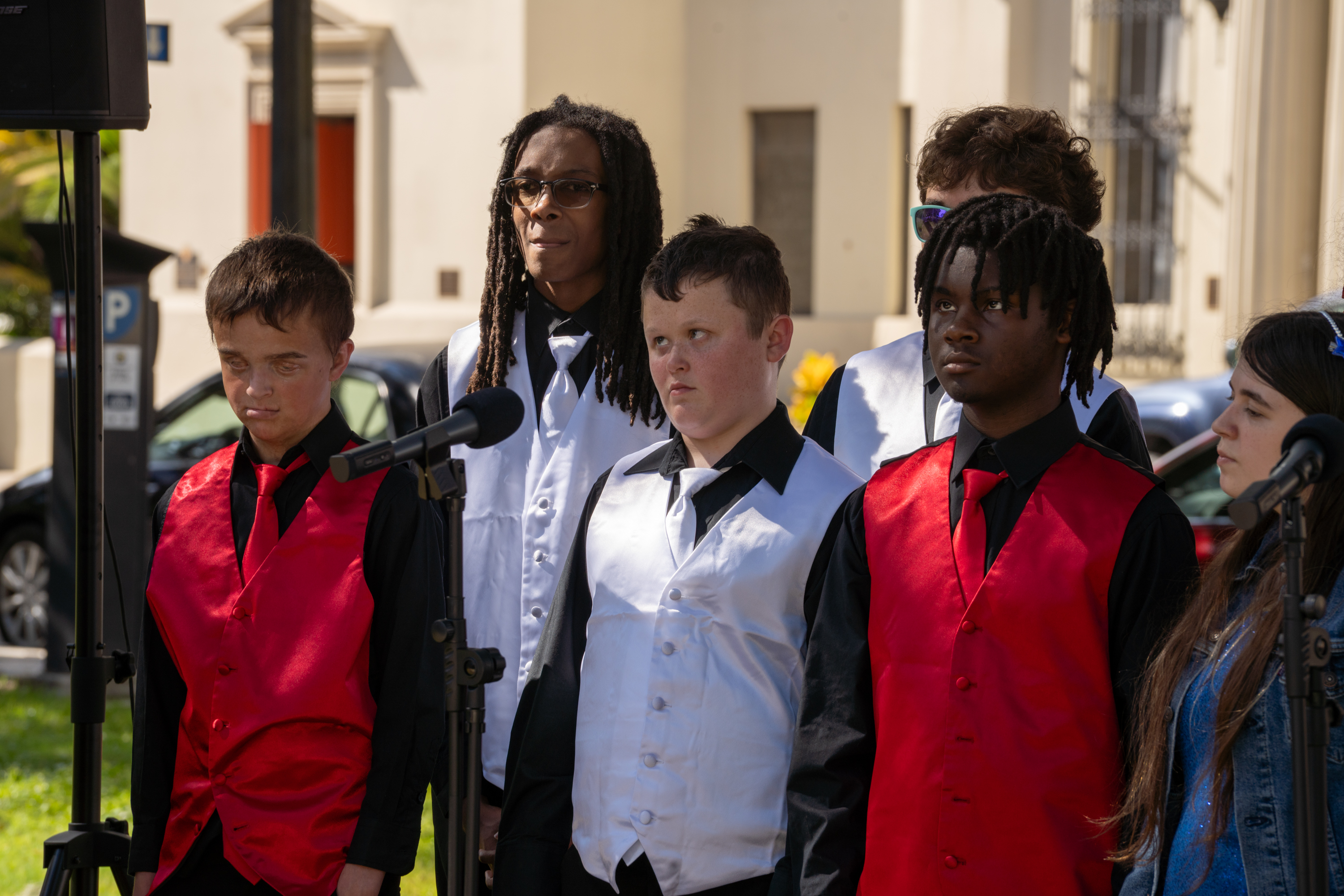 The boys in the group are standing in concert position. 3 of the boys are wearing white vests and ties while 2 of the boys are wearing red vests and ties.