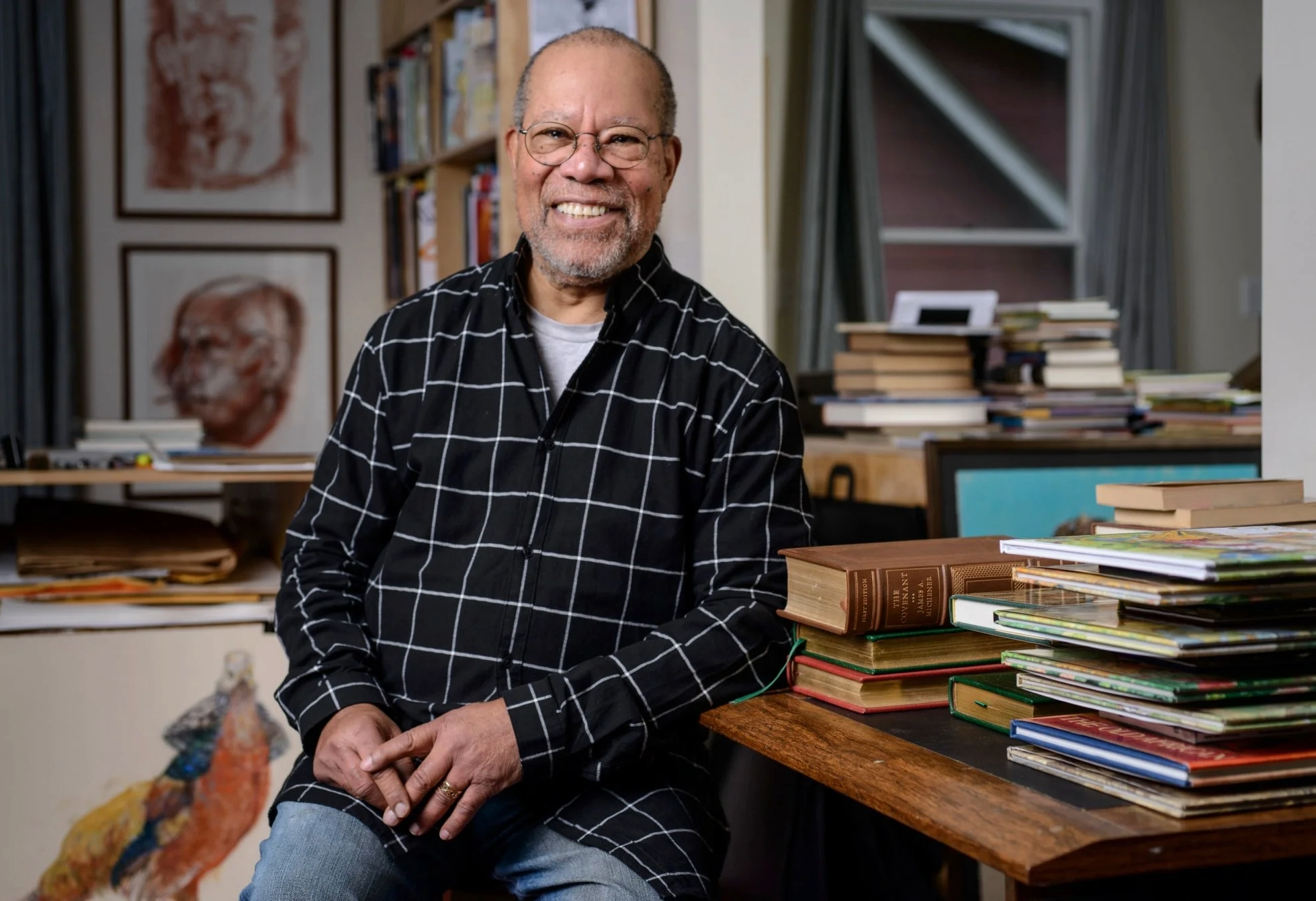 Jerry Pinkney smiling shile leaning against a table with several stacks of books