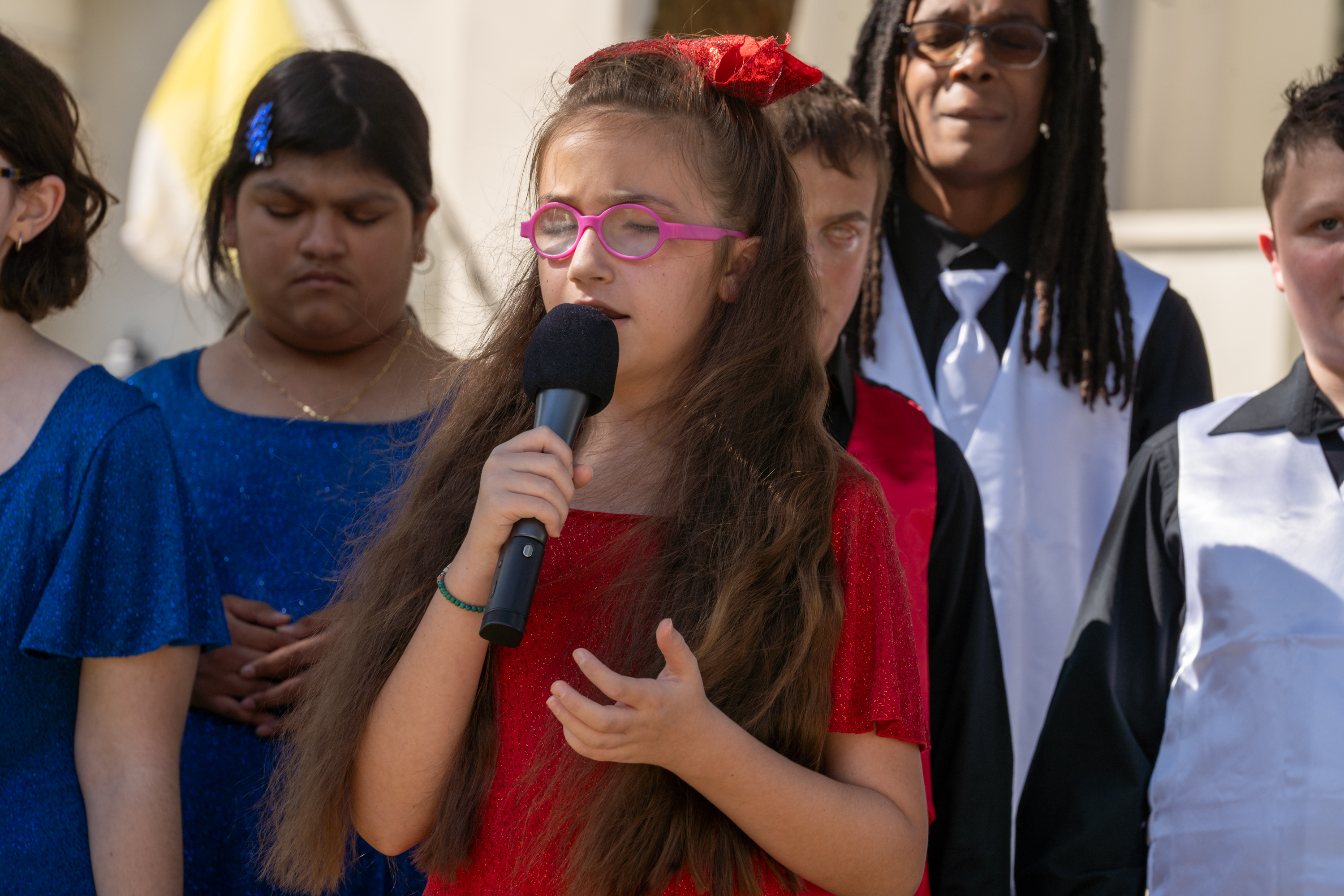 McKenzie singing a solo. She is wearing a red glitter dress and has a red sequin cheer bow in her hair. she is holding a microphone in her right hand and her left hand is held up facing sky in a gesture.