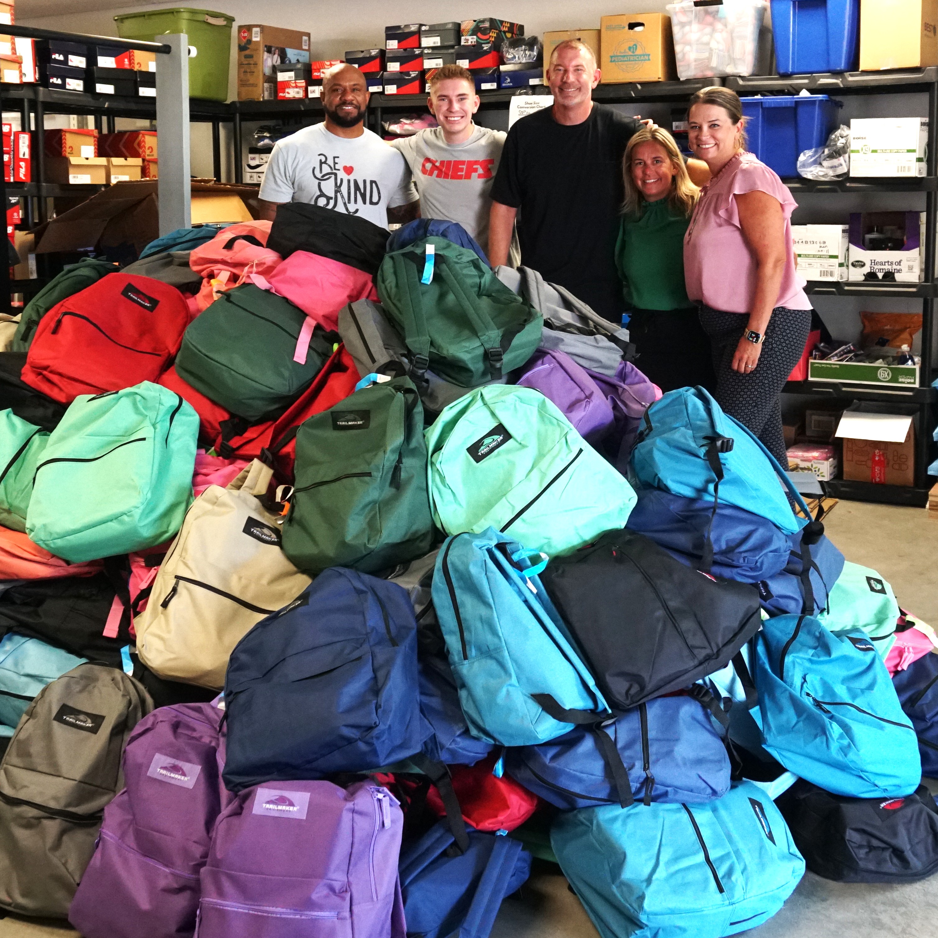 Organizers and donors stand arm in arm behind a mountain of backpacks stuffed with school supplies