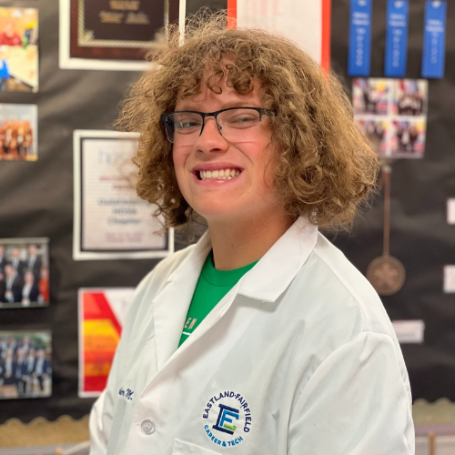 Alexander Mayeres, a Bioscience senior, smiles while wearing glasses and a white Eastland-Fairfield lab coat, standing in front of a bulletin board with awards and photos.