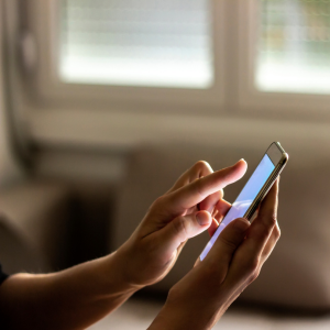  Close-up of a person's hands holding and using a smartphone, with their finger tapping on the screen. The background is blurred, showing a window with blinds.