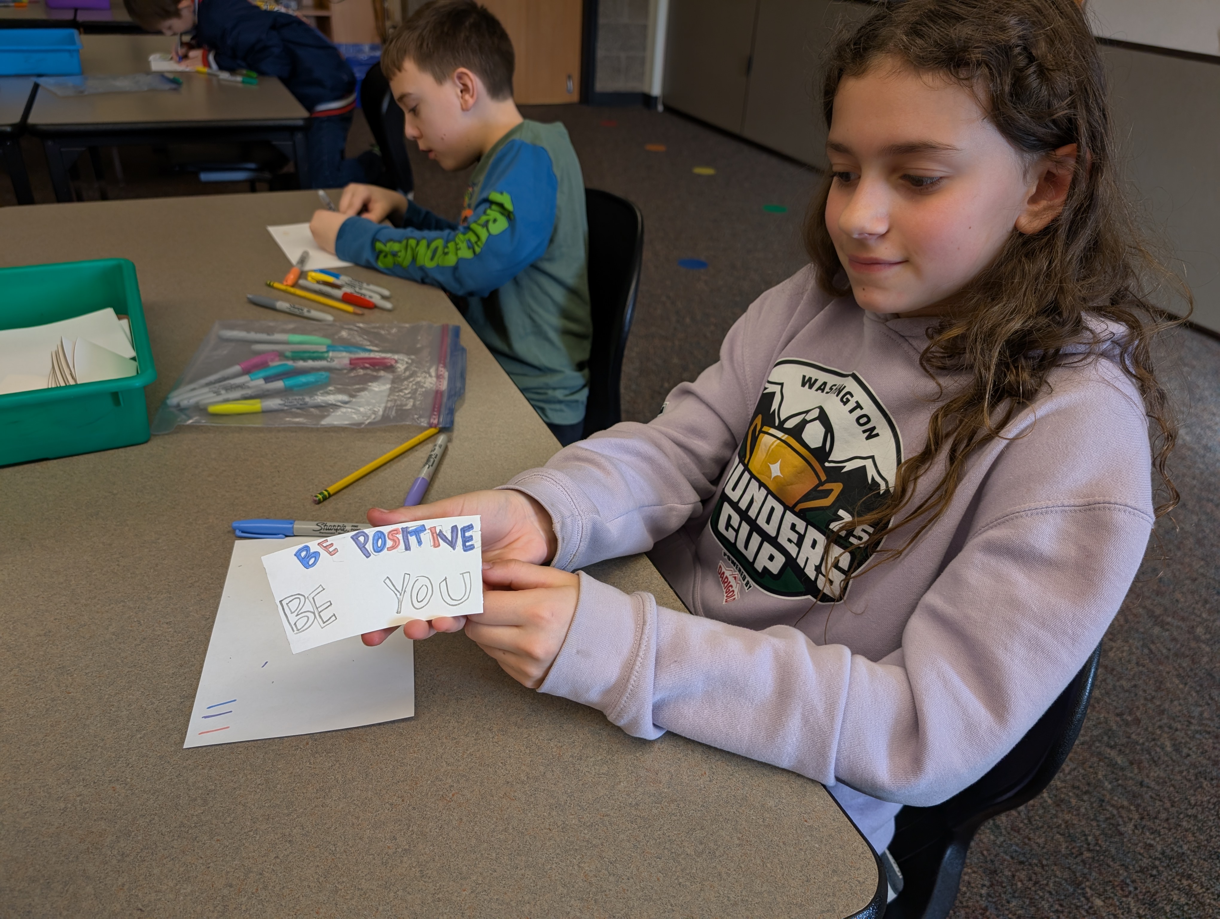 A student holds up a coffee cup sleeve on which she is writing the words "be positive, be you"