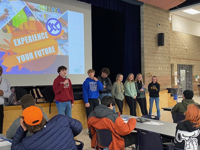 students standing in front of a stage with a screen behind them that says "Experience Your Future SCC" and students in front of them  listening to a presentation