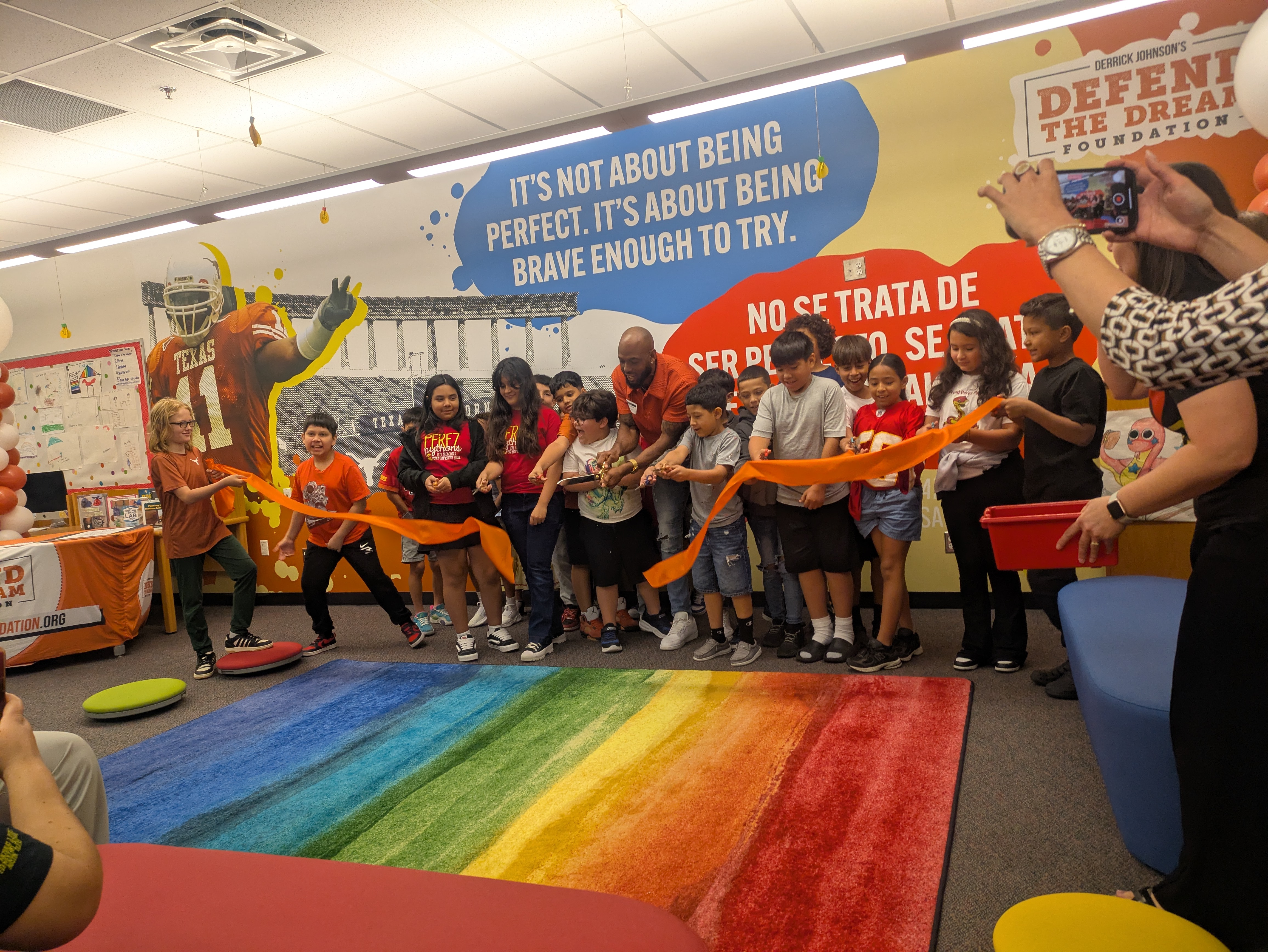 A group of elementary students and adults participate in a ribbon-cutting ceremony inside a colorful classroom with a rainbow rug. The wall behind them features a large mural of a Texas Longhorns football player and motivational quotes in English and Spanish. The event is part of the grand opening of DJ’s Discovery Den, supported by Derrick Johnson’s Defend the Dream Foundation. A few attendees capture the moment on their phones.