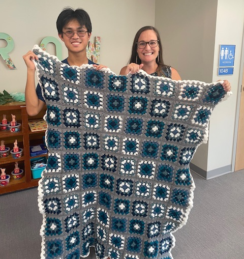 Gabe and a teacher holding up a gray, black, white, and navy granny square blanket
