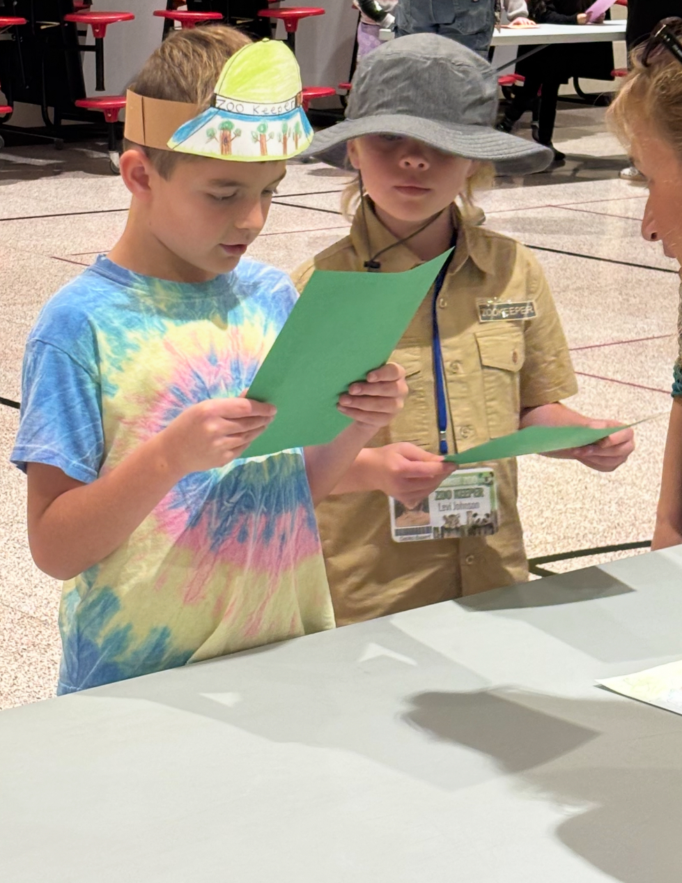 Two students with hats holding green papers and reading to an adult.