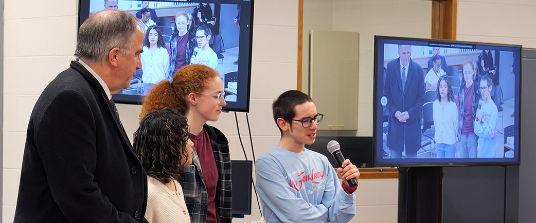 Student talking into microphone while  two other students and an adult look on.