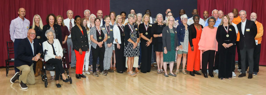 A large group of men and women stand together on a gymnasium floor in front of maroon curtains and a black backdrop. Many are wearing medals around their necks, and several individuals are dressed in Tennessee High’s colors of maroon, black, and orange. One woman is seated at the front, and others stand in rows behind her, smiling for the photo. The group represents the Bristol Tennessee City Schools Athletic Hall of Fame Class of 2025 during their induction event at Tennessee High’s Viking Hall.
