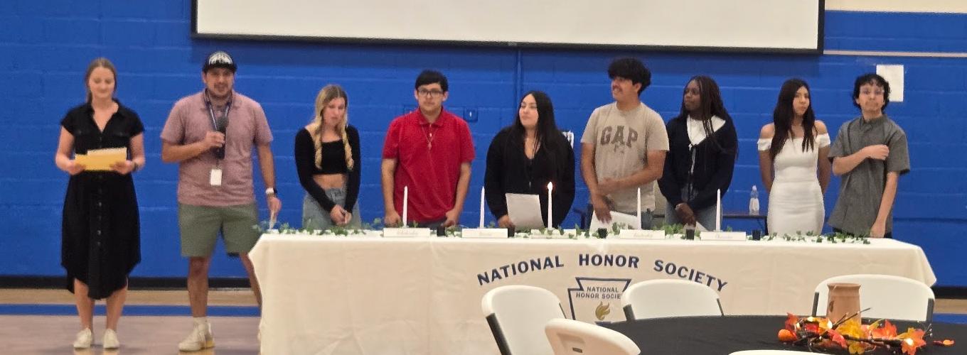 Students and teachers standing behind a table with a "National Honor Society" tablecloth and candles.
