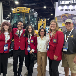 A group of five students in red SkillsUSA jackets and their advisor, standing in front of a Caterpillar construction vehicle at a conference hall. The background includes a large Caterpillar banner.