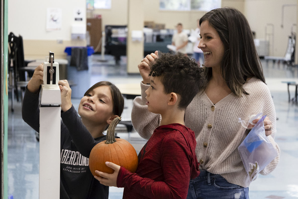 A parent helping two students weigh a pumpkin