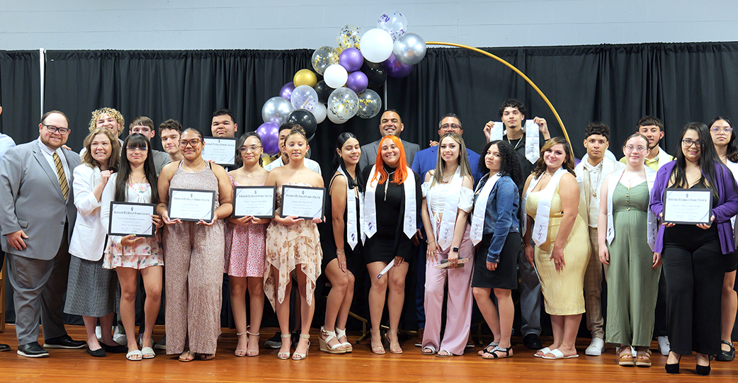 Group of Dean students holding certificates, and wearing stoles, on stage with staff 