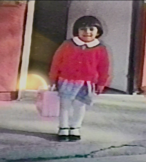 Guess Who: Pictured is a student in a cherry red sweater, blue skirt, white tights and black Mary Jane shoes, holding a pale pink lunchbox.