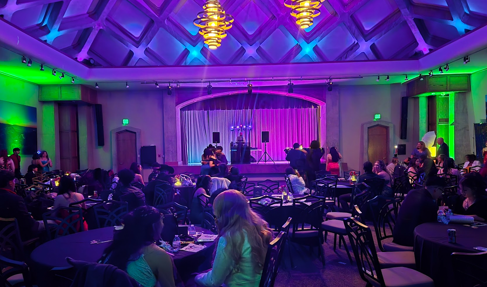 Low lit prom venue with students sitting at tables and a stage at the front of the room