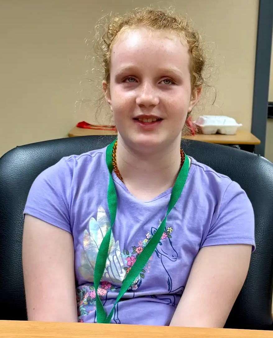 Maely. She is wearing a lavender t-shirt and has her green school lanyard around her neck. Her curly ginger hair is pulled back in a ponytail.