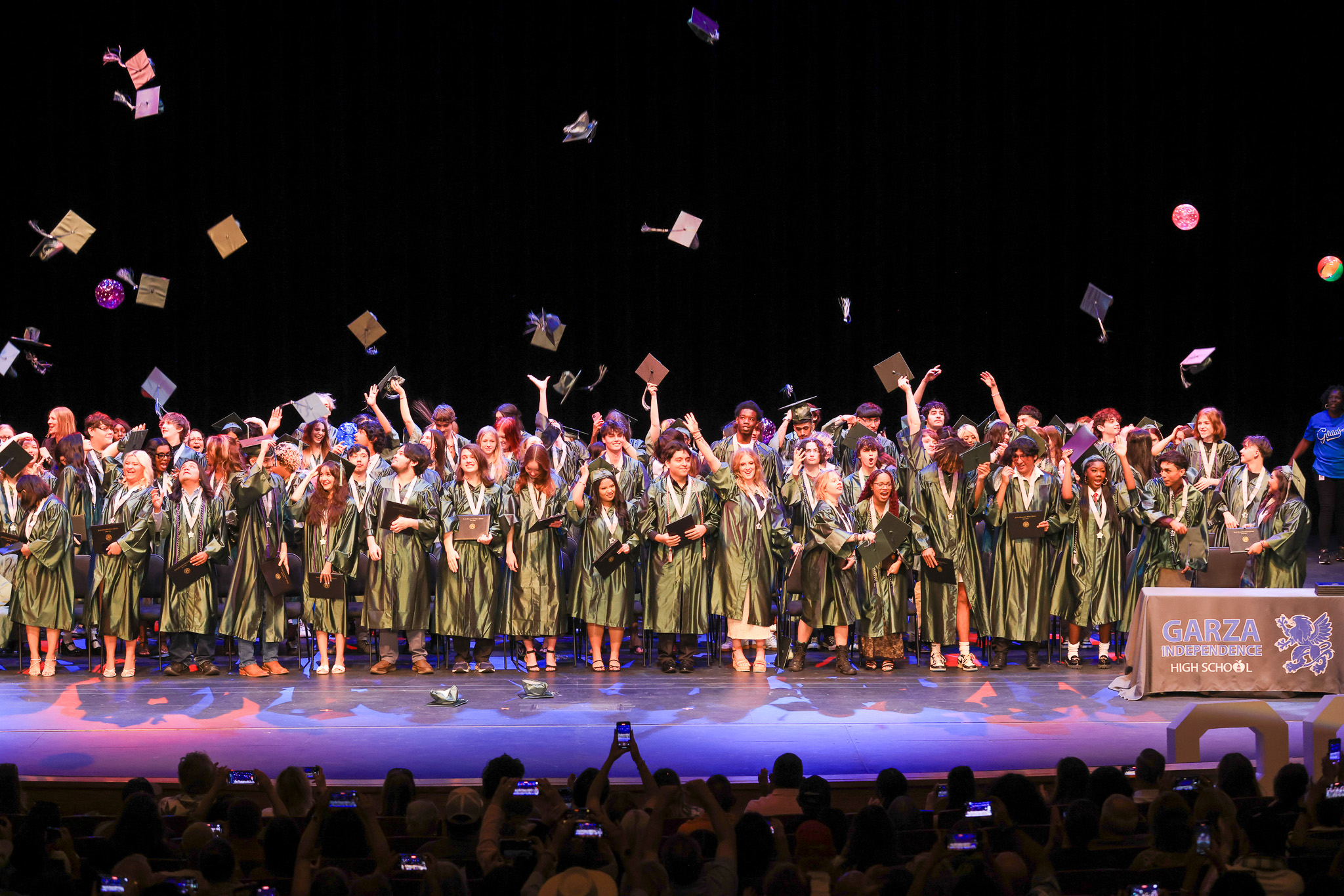 A large group of graduating students from Garza Independence High School stand on stage in green caps and gowns, cheering and tossing their caps into the air. Many hold diplomas and medals, and the audience below captures the moment on their phones. A table on the right displays the school name and logo.