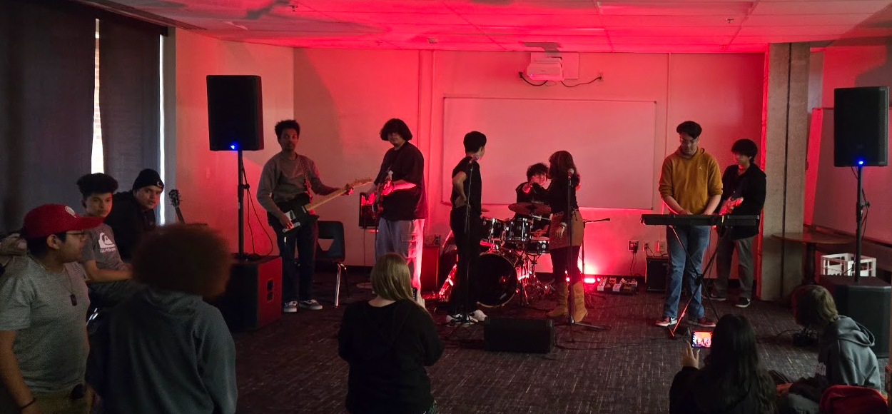 Students playing keyboard, guitar and drums at the front of a low-lit, red room
